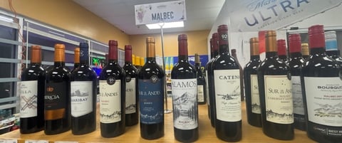 Row of red wine bottles displayed on a shelf in a wine shop with store signage visible in the background