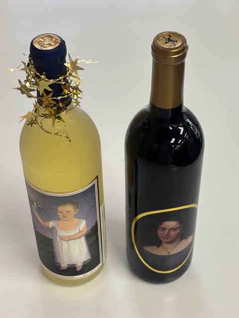 Two decorative wine bottles with personalized labels and portrait images, one with yellow wine and gold ornament bow, one with red wine and bronze cap