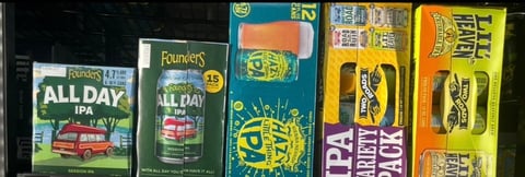 Collection of craft beer cans and boxes displayed on shelf, including Founders All Day IPA and various other colorful brewery packaging