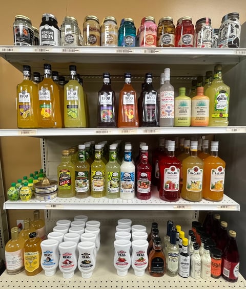 Retail shelf display of alcoholic beverages including beer cans, liquor bottles, and mixers organized on four white shelves